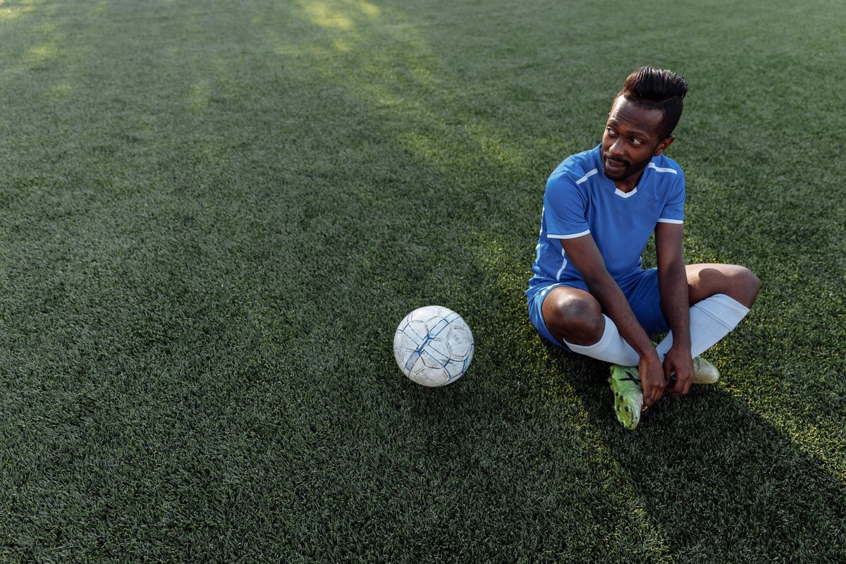 Footballer in a blue football shirt sitting on the pitch beside a match ball